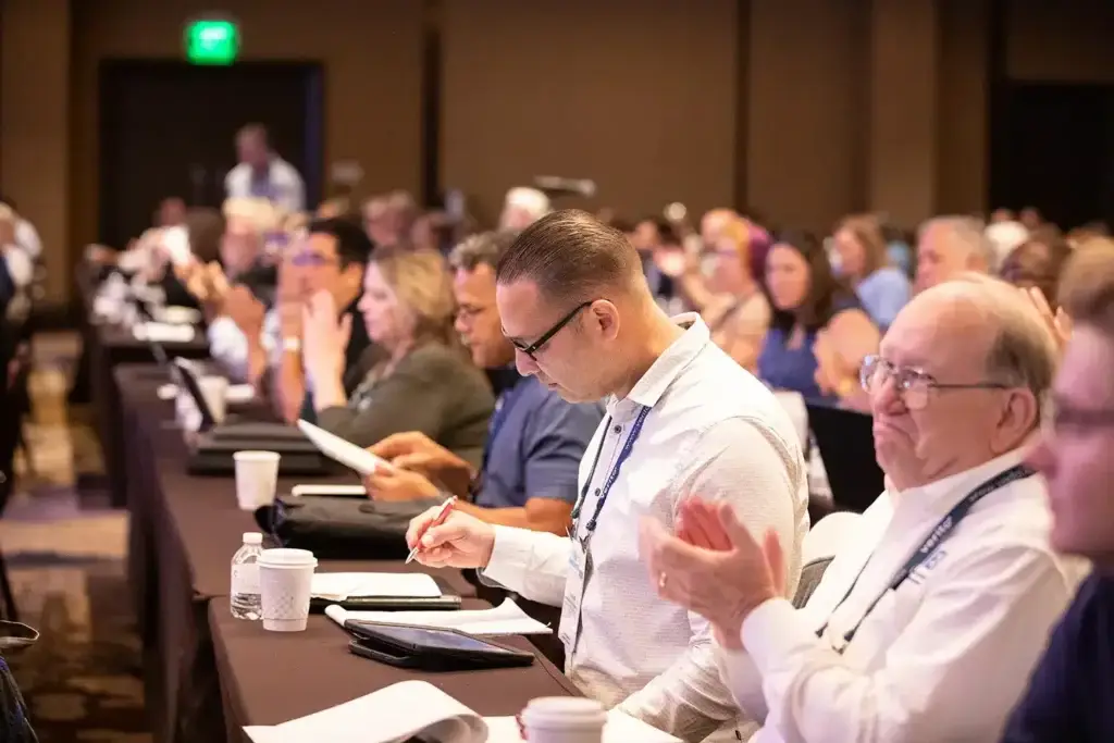 Conference attendees seated at a long table, a man in a white shirt takes notes while others applaud in the background.