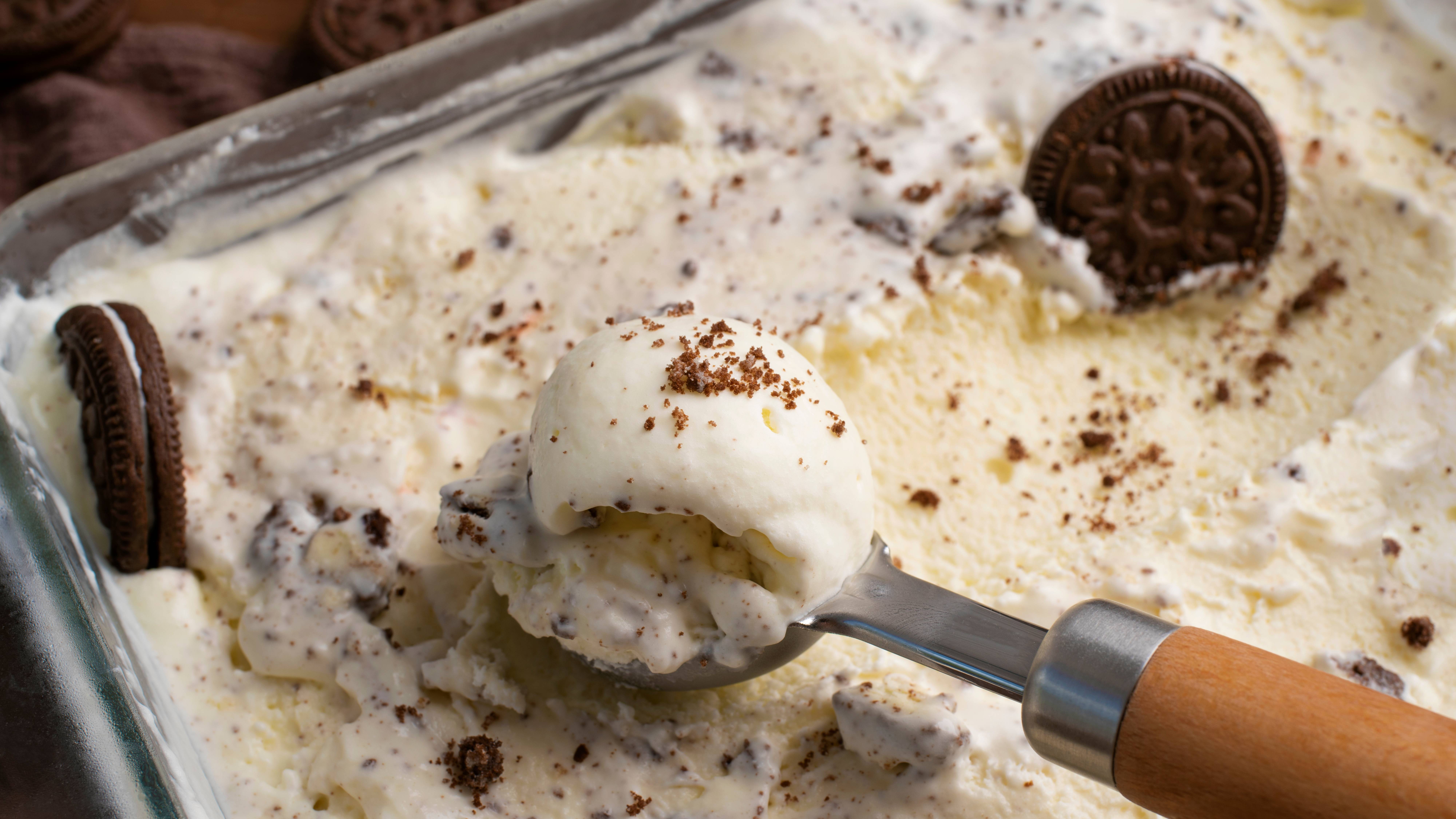 Scoop of cookies and cream ice cream with Oreo cookie pieces and a whole Oreo nearby on a dish, with a metal ice cream scoop in view.